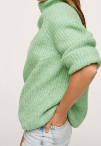 Pull tricoté vert menthe avec un col haut, des manches retroussées et un motif texturé. Porté sur un jean bleu clair, présentant une coupe ample.