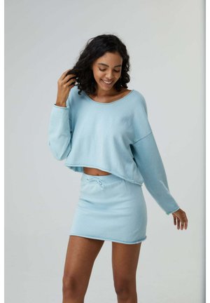 Lächelnde Frau mit lockigem Haar trägt hellblauen langärmeligen Crop-Pullover und passenden Minirock mit hoher Taille vor einfarbigem Hintergrund.