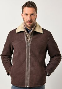 Hombre con una chaqueta de ante marrón y cuello de borrego beige, vestido con varias capas de prendas de punto, de pie con las manos en los bolsillos frente a un fondo sencillo.
