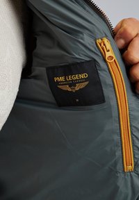 Giacca grigia isolata con zip arancione e etichetta nera con testo dorato che recita "PME LEGEND AMERICAN CLASSIC," taglia M.