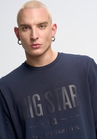 Jūras zilas krāsas svārki, kas izgatavoti no mīkstas auduma, ar lielu reljefu uzrakstu "BIG STAR" un papildu tekstu apakšā. Apaļais kakls, faktūru detaļas.