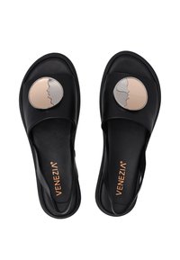 Schwarze Slipper-Sandalen mit einem breiten Obermaterial, das einen runden dekorativen Akzent aus zwei verschlungenen Gesichtern in beige und silbernen Tönen zeigt.