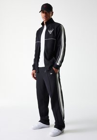 Chándal negro con franjas laterales blancas, chaqueta de cremallera con logo, combinado con pantalones a juego y zapatillas blancas. Gorra negra con logo.