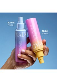Main tenant deux sprays fixateurs IsaDora, un fini mat violet et un fini lumineux rose et jaune, sur fond bleu.