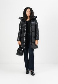 Zwarte, glanzende gewatteerde jas met een capuchon, lang model, met een gestept ontwerp. Gepaard met donkere denim broeken en een kleine zwarte tas.