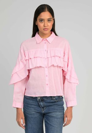 Femme portant un chemisier rose clair à boutons avec des volants superposés sur la poitrine et les manches, assorti à un jean bleu, sur un fond uni.