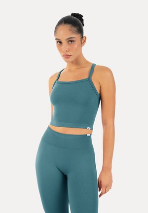 Kvinde med håret bundet tilbage iført en teal tætsiddende cropped top og højtaljede leggings foran en ensfarvet baggrund, kigger fremad med et neutralt udtryk.