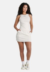 Abito bianco aderente con design a canotta, dotato di tasche laterali e cuciture a contrasto. Il modello indossa sneakers bianche e calzini alla caviglia.