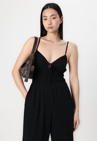 Macacão preto com alças finas, decote em V profundo e cintura com cordão ajustável. Modelo segura uma pequena bolsa de couro castanho com uma alça ajustável.