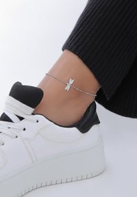 Sølv ankelkæde med en guldsmed vedhæng, båret med en sort ribbet tekstil og en hvid sneaker med sorte accenter.