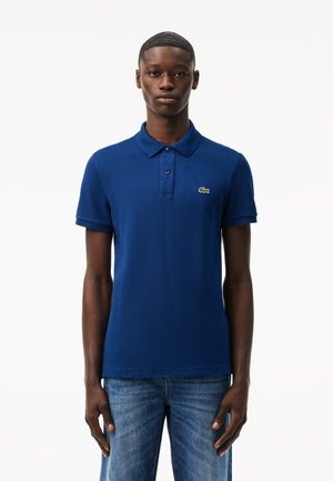 Marinblå piké med en texturerad tyg, treknappsknäppning och en liten grön krokodil-logotyp på vänster bröst. Bärs tillsammans med blå jeans.