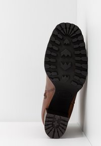 Botte marron positionnée semelle vers le haut contre un mur d'angle blanc, montrant une épaisse semelle noire à crampons avec des rainures profondes pour l'adhérence.