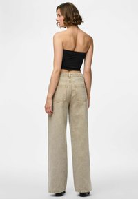Donna con capelli corti e mossi, che indossa un top nero senza spalline e jeans beigi alti in vita e a gamba larga, in piedi su uno sfondo bianco.