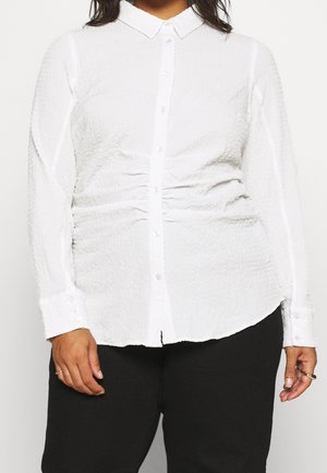 Chemise blanche à manches longues texturée avec des boutons et un détail froncé à la taille, portée avec un pantalon noir, montrant des mains avec des bagues.