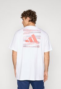 Hvit bomulls t-skjorte med et stort rosa og grått abstrakt Adidas-logo på ryggen. Korte ermer og en avslappet passform.