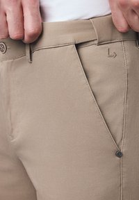 Beżowe bawełniane spodnie z teksturowanej powierzchni, z guzikiem i subtelnym logo na pasku. Wyraziste zagniecenia podkreślają design.