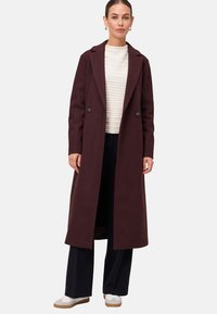 Vrouw in een lange donkerbruine jas over een gestructureerde witte top, zwarte wijduitlopende broek en witte sneakers, staande tegen een effen achtergrond.