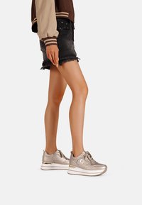 Sneakers metallici dorati con una finitura liscia, chiusura con lacci e suola bianca; indossati con una gonna nera a orlo sfrangiato e una giacca bomber marrone.