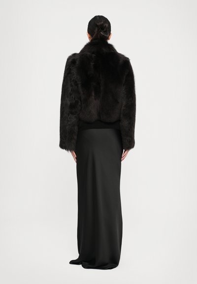 Jachetă scurtă din blană sintetică neagră, cu mâneci largi, asortată cu o fustă lungă din satin negru. Textură netedă și design simplu, vedere din spate.