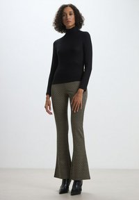 Un pull noir à col roulé côtelé associé à un pantalon évasé noir et doré à motifs. Le mannequin porte des bottines noires, posant devant un fond neutre.