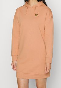 Vestido sudadera con capucha en melocotón, fabricado en tela suave. Presenta mangas largas, un corte holgado y un pequeño logo de un pájaro amarillo en el pecho.