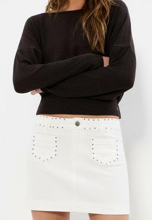 Mujer vistiendo una camiseta negra de manga larga y una minifalda blanca con detalles de tachuelas plateadas en los bolsillos y la cintura.