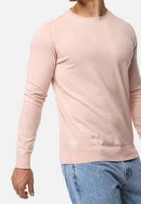 Pull à col rond rose clair avec manches longues, poignets et ourlet côtelés. Matière douce avec une texture lisse. Porté avec un jean denim bleu.