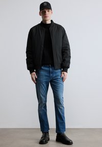 Μαύρο μπουφάν bomber, πλεκτό μαύρο πουλόβερ, μπλε τζιν και μαύρα παπούτσια, συμπληρωμένα με μαύρο καπέλο με λευκό κείμενο. Casual αστικός στιλ.