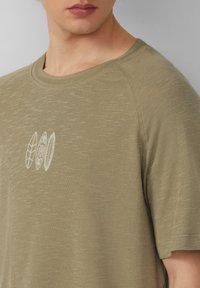 Oliwkowa koszulka z okrągłym dekoltem, uszyta z miękkiego materiału, z textured wykończeniem i białymi haftowanymi wzorami trzech liści po lewej stronie.