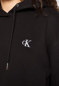 Personne portant un sweat à capuche noir avec le logo blanc "cK" brodé sur la poitrine, cheveux foncés partiellement visibles.
