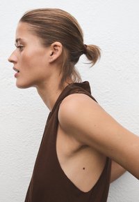 Haut marron sans manches avec une large découpe aux aisselles laissant la peau visible. Cheveux attachés en un chignon bas. Fond clair avec un mur texturé.