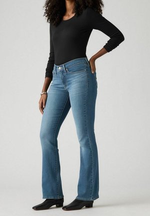 Femme portant un haut noir moulant à manches longues, un jean bootcut bleu clair et des bottines noires, debout avec une main sur la hanche.