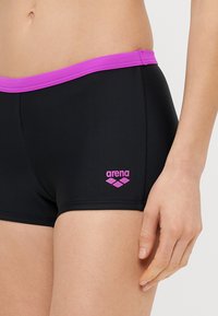 Κοντινό πλάνο ενός ατόμου που φοράει μαύρο μαγιό με μοβ ζώνη μέσης και το λογότυπο Arena στο αριστερό μηρό, με το χέρι να ακουμπά στο πλάι.