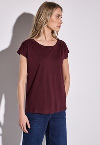 Top burdeos de manga corta con escote redondeado y detalles de hombros fruncidos, combinado con pantalones de mezclilla azul. Textura suave, ajuste relajado.