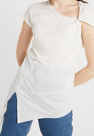 Haut beige clair à manches courtes avec un détail noué à l'épaule. Confectionné en tissu doux, il présente un ourlet asymétrique et une surface texturée.