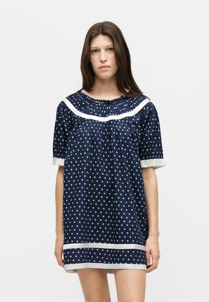 Fiatal nő egyenes barna hajjal sötétkék, rövid ujjú ruhát visel, fehér pöttyökkel és fehér szegéllyel az ujjakon és az alján.