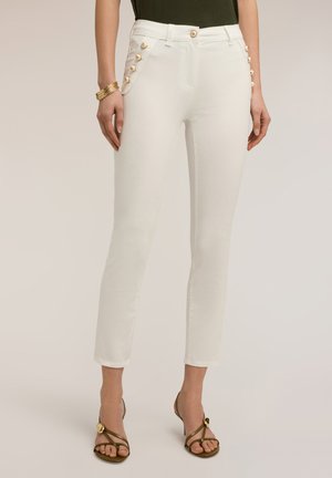 Vrouw draagt witte slim-fit broek met gouden knopen op de zakken, gecombineerd met een olijfgroene top, gouden armband en bruin open-toe hakken.