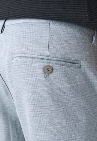 Gros plan sur la poche d'un pantalon gris clair texturé avec un seul bouton, porté avec un pull noir côtelé.
