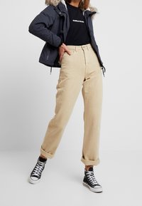 Beige høj talje bukser med en løs pasform, foldede manchetter, parret med en sort skjorte og en marineblå pufferjakke med en faux fur hætte.