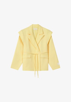 Blazer sur mesure jaune clair avec épaules rembourrées, fermeture à bouton unique, poches avant et ceinture nouée à la taille sur fond blanc.