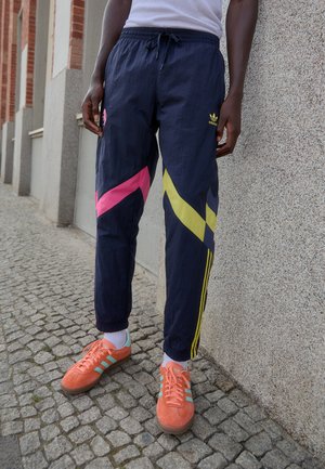 Navy Jogginghosen mit pinken und gelben diagonalen Streifen, elastischem Bund, seitlichen Streifen und einem gestickten Logo. Orangefarbene Sneakers mit türkisfarbenen Akzenten.