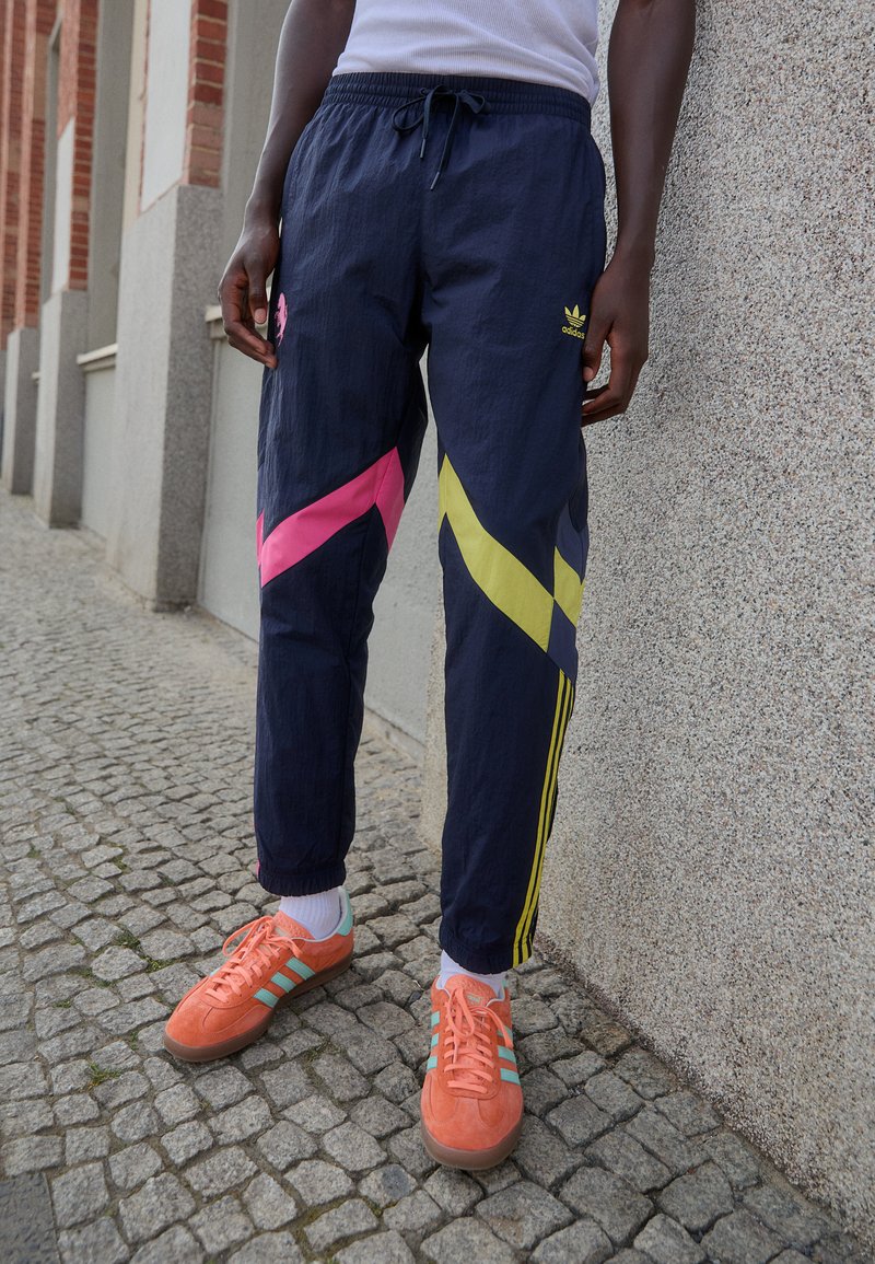 Pantaloni da jogging navy con strisce diagonali rosa e gialle, vita elasticizzata, strisce laterali e logo ricamato. Sneakers arancioni con dettagli in teal.