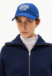 Person, der eine blaue Kappe mit dem Schriftzug "CHANGE MAKERS CLUB", einen marineblauen Reißverschluss-Pullover über einem weißen, kabelgestrickten Rollkragenpullover trägt und nach vorne schaut.