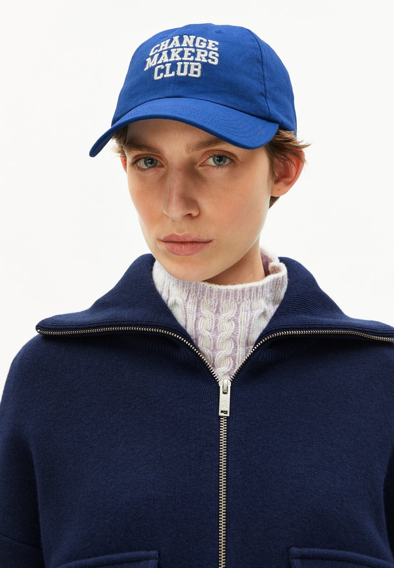 Person, der eine blaue Kappe mit dem Schriftzug "CHANGE MAKERS CLUB", einen marineblauen Reißverschluss-Pullover über einem weißen, kabelgestrickten Rollkragenpullover trägt und nach vorne schaut.