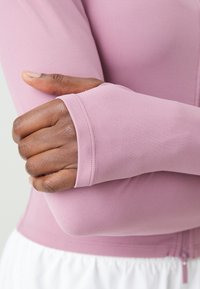 Dlouhý rukáv top v měkké, elastické růžové tkanině s přiléhavými manžetami. Disponuje zipovým zapínáním vpředu a hladkým povrchem, vhodný na sportovní oblečení.