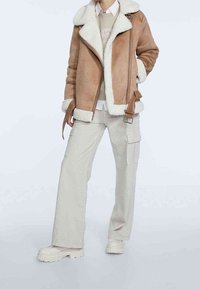 Persoon in een beige schapenvachtjack met riem, lichte cargo-broek en stevige off-white laarzen tegen een effen achtergrond.