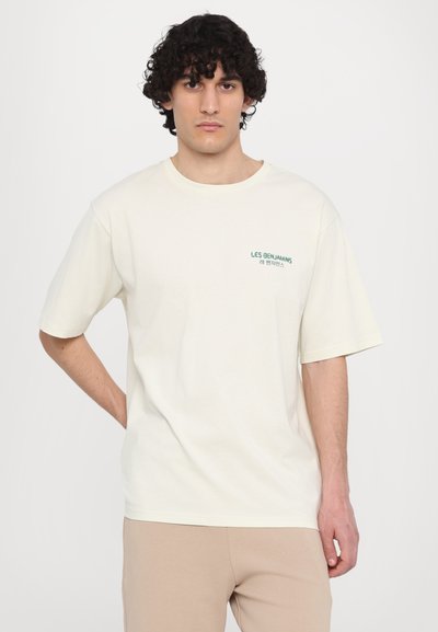 T-shirt crème à manches courtes en coton doux, arborant un petit logo textuel graphique vert sur la poitrine gauche. Coupe décontractée, col rond.