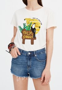 Kermainen puuvillainen t-paita, jossa värikäs tukaani- ja papukaijakuvio sekä "PALM STREET" -kyltti, yhdistetty rispaantuneisiin farkkushortseihin, joissa viisipokkainen muotoilu.