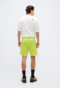 Άνδρας που μοιάζει από πίσω, φοράει λευκό μπλουζάκι North Face, φωτεινό κίτρινο σορτς, μαύρες κάλτσες και μαύρα αθλητικά παπούτσια Salomon, σε απλό λευκό φόντο.