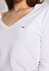 Valkoinen pitkähihainen V-päätteinen toppi pehmeästä kankaasta, jossa on pieni brodeerattu logo punaisena, valkoisena ja sinisenä vasemmassa rinnassa.